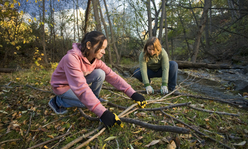 Two girls picking up sticks in a wooded area.